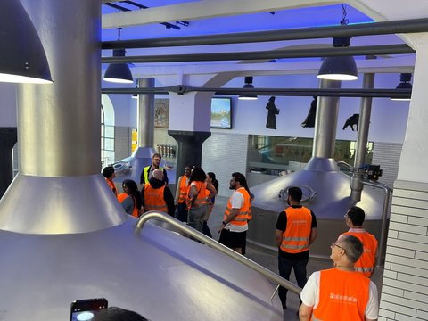 Die Seminargruppe in orangefarbenen Warnwesten hört einem Mitarbeiter im Sudhaus der Brauerei Weihenstephan zu. Große silberne Braukessel umgeben die Gruppe.