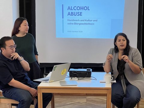 Die drei Geförderten während der Präsentation zum Alkoholmissbrauch. Adriana spricht und gestikuliert; hinter ihnen ist die Titelfolie zu sehen.